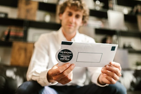 A man sitting on a couch holds an envelope marked with "Pre-Qualified" and "Unlimited 1.5% Cash Back."