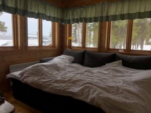 **Alt text:** A cozy bed with white linens is placed in front of large windows in a wooden cabin, overlooking a snowy landscape.