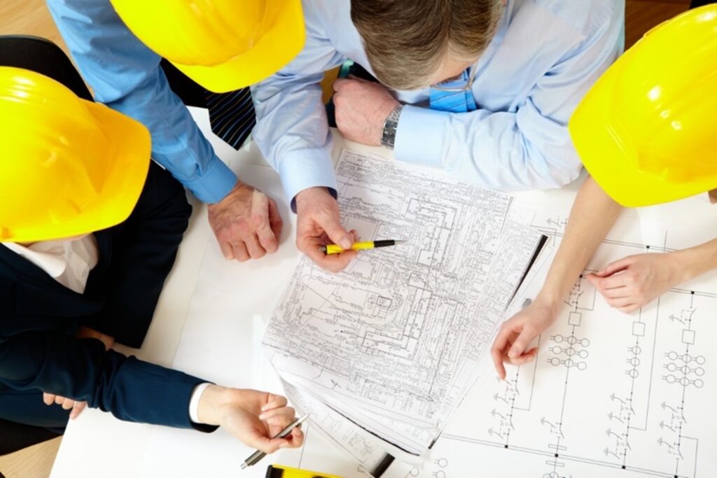 **Alt Text:** A group of people wearing yellow hard hats and blue shirts are gathered around a table, examining architectural blueprints.