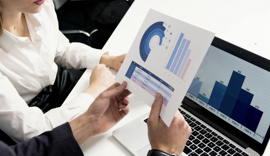 **Alt Text:** Two people in a business setting analyzing graphs on a printed document and a laptop screen.