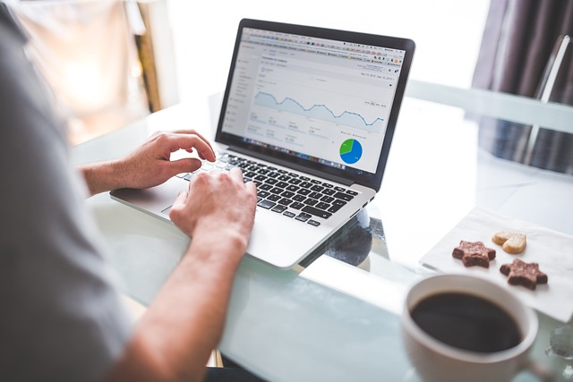 **Alt text:** A person working on a laptop displaying data graphs, with a cup of coffee and cookies on the table.