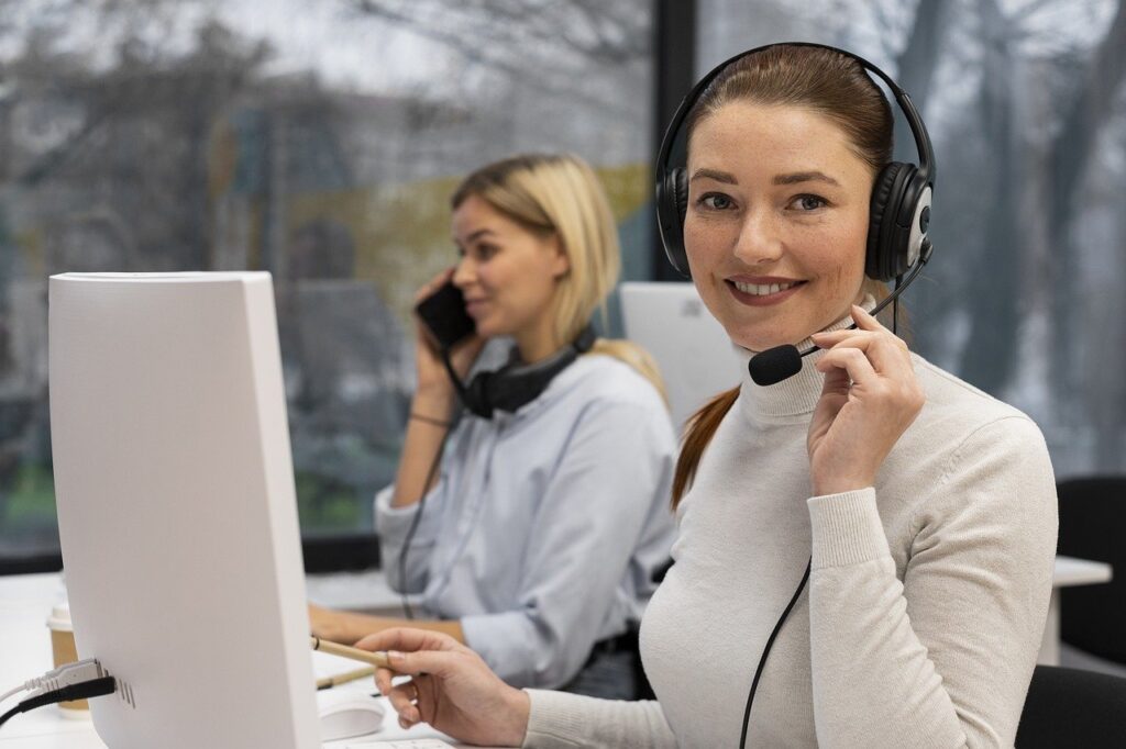 Two women in an office setting wearing headsets, with one smiling at the camera and the other talking on the phone.