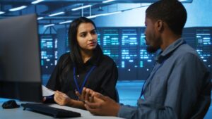 **Alt Text:** Two professionals engaged in discussion seated in a modern data center with glowing servers in the background.