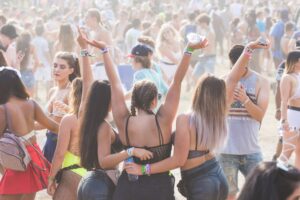 **Alt text:** A group of people enjoying a lively outdoor festival, with three women in the foreground in stylish outfits, raising their arms and cheering amidst a crowd.