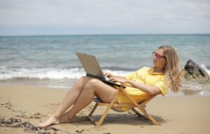 A person in a yellow shirt and red glasses is sitting in a beach chair using a laptop by the sea.