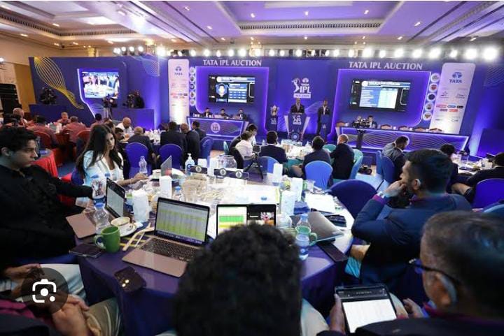 **Alt Text:** A large room filled with people seated at round tables, equipped with laptops and tablets, attending a cricket player auction event with a blue-lit stage at the front displaying the "Tata IPL Auction" banner.