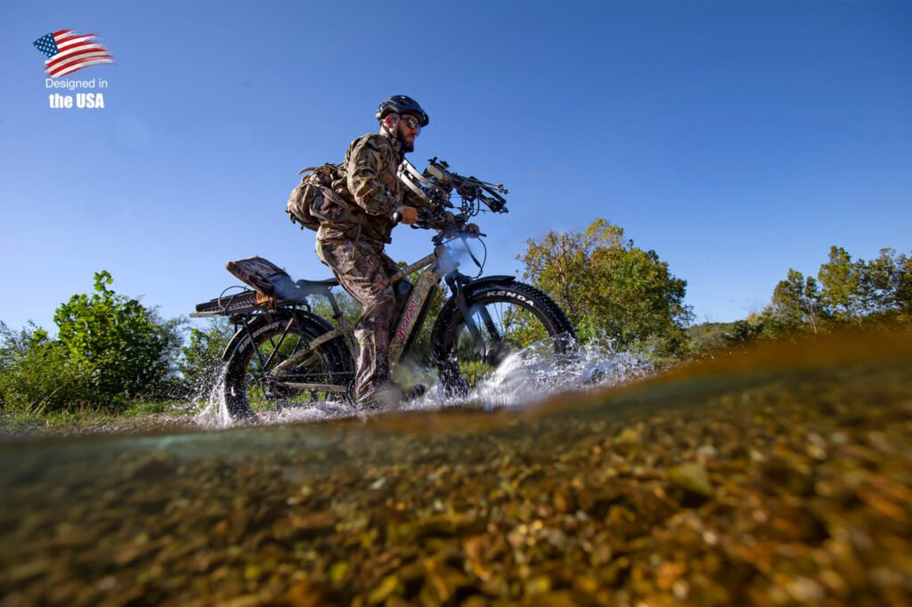 A person in camouflage gear rides a rugged bike through shallow water with trees and a clear blue sky in the background.