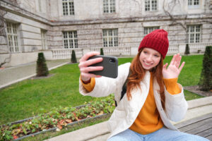 A person in a white coat and red beanie takes a selfie while sitting on a bench in a courtyard.