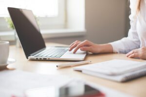 A person using a laptop on a wooden desk with a notebook, pen, and coffee cup nearby.
