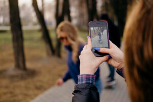 A person holds a smartphone, capturing a photo of a woman skateboarding on a path through a park.