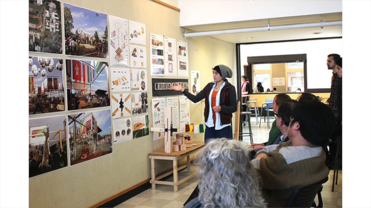 A person presents architectural design concepts on a wall to a seated audience in a classroom setting.