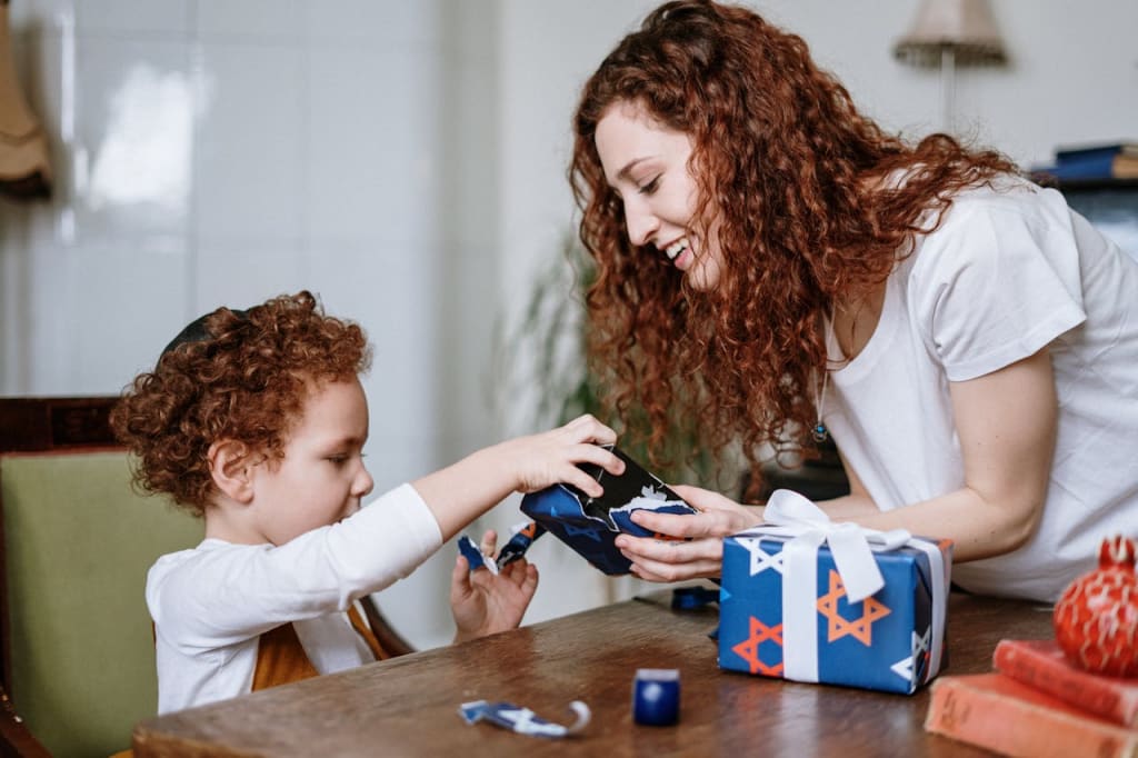 A woman and a child with curly hair happily wrapping a gift with Star of David patterned paper at a table.