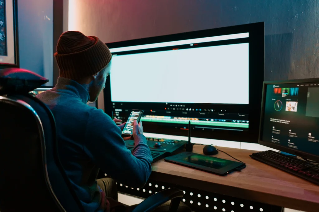 A person wearing a beanie and sweater is sitting in a dark room, looking at a smartphone in front of multiple computer screens displaying editing software.