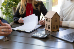 Three people sitting at a table reviewing documents with a model house, calculator, and laptop in view.