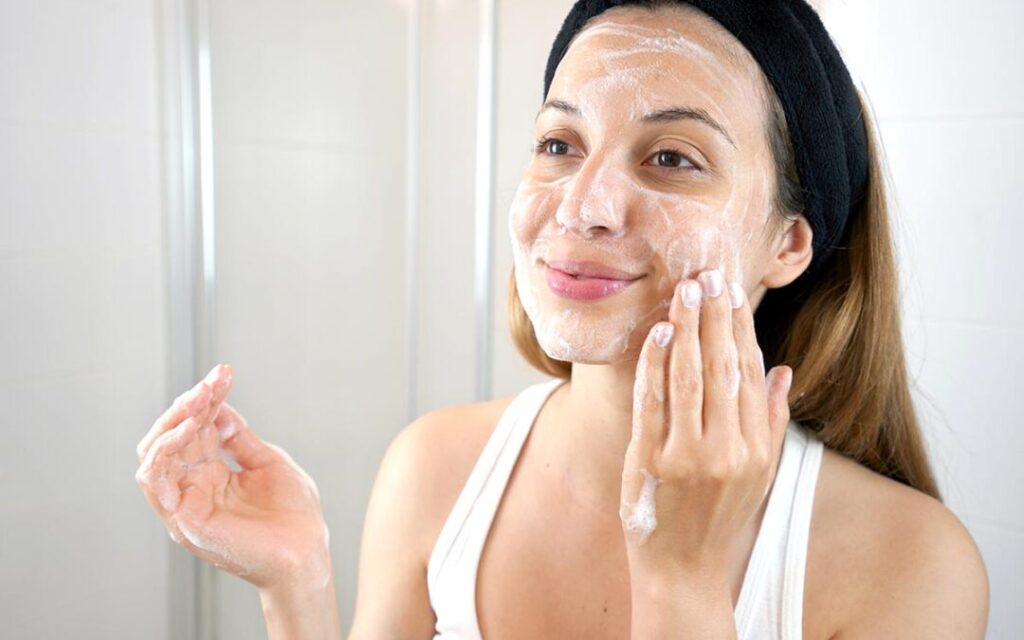 A woman in a bathroom applies a foaming facial cleanser to her face.