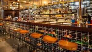 **Alt text:** A modern bar with a long counter, high wooden stools, and an array of bottles and glasses on shelves, featuring a playful design with colorful Lego bricks incorporated into the base structure.