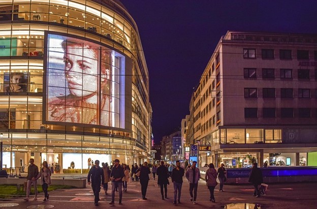 **Alt Text:** A busy city street at night with people walking past a large building displaying a digital advertisement featuring a woman's face.