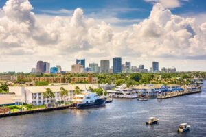 A waterfront cityscape with modern buildings, palm trees, yachts, and boats under a partly cloudy sky.