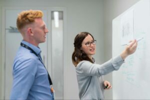 A woman writing on a whiteboard while a man observes in an office setting.