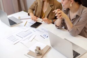 Two people are seated at a table with laptops, reviewing printed financial documents and using a calculator.