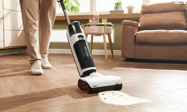 A person uses a vacuum cleaner to clean a spill of cereal and milk on a hardwood floor in a living room.