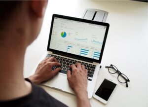 **Alt text:** A person is using a laptop to view data analytics with graphs and charts, with a smartphone and glasses on the desk beside them.