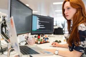 A woman with red hair is focused on coding at a dual-monitor setup in an office environment.