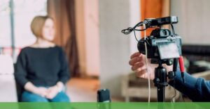 **Alt text:** A camera on a tripod is focused on recording a person sitting in a chair during an indoor interview setup.