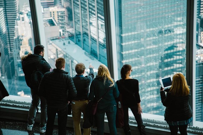 **Alt Text:** A group of people stand by large windows in a skyscraper, gazing out over the city below.
