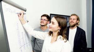 A woman writes on a flipchart while two men watch attentively in a professional setting.