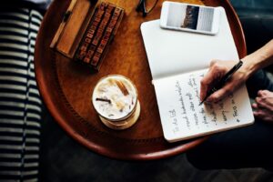 A person writes in a notebook with a to-do list on a round wooden table, next to an iced coffee, a smartphone, and an abacus.