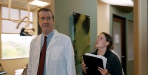 A doctor in a white coat walks through a hallway with a woman carrying a clipboard beside him.
