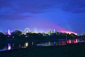 **Alt Text:** A vibrant night scene featuring an amusement park with a brightly lit Ferris wheel and other illuminated rides, reflected in a calm river.