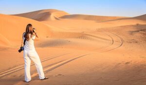 **Alt text:** A woman in white clothing stands on sand dunes with a camera in hand, looking out at tire tracks and undulating desert landscape under a clear blue sky.