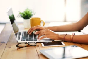 **Alt text:** A person typing on a laptop at a wooden table with glasses, a smartphone, a tablet, and a yellow mug nearby.