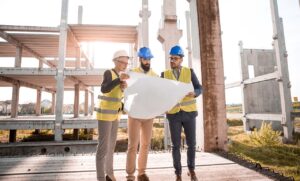 Three construction workers wearing hard hats and reflective vests examine a blueprint at a building site.