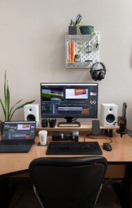 A home office setup with a desktop monitor, laptop, speakers, camera equipment, and a wall shelf holding books and plants.