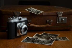 A vintage camera is placed on a wooden table next to scattered black-and-white photographs and in front of an old brown suitcase.