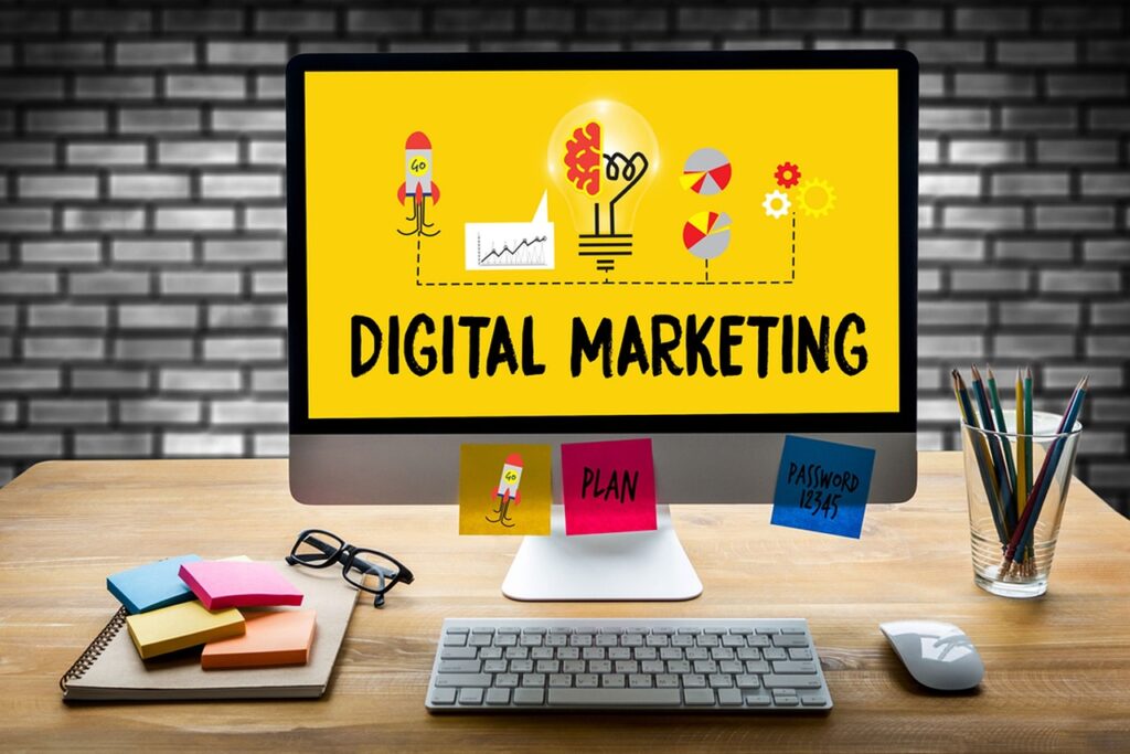 A computer screen displaying "Digital Marketing" with graphics of a rocket, light bulb, and pie charts on a yellow background, on a desk with colorful sticky notes, a keyboard, and pens.