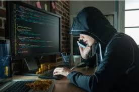 A person wearing a hoodie sits in front of a computer with code on the screen, talking on a smartphone, in a dimly lit room.
