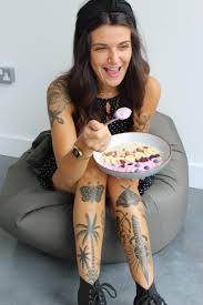 A person with long hair and tattoos on their arms and legs is sitting on a bean bag, holding a bowl of cereal and smiling.