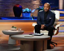 **Alt Text:** A person in a suit is seated on a modern chair in a well-lit studio setting with a round table beside them.