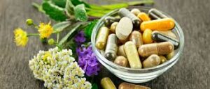 **Alt Text:** A small glass bowl filled with various colored capsules and tablets is surrounded by assorted fresh herbs and flowers on a wooden surface.