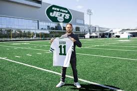A person stands on a football field holding a New York Jets jersey with the name "Dior" and the number 11, in front of the Jets training facility.