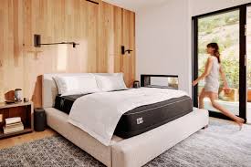 A modern bedroom with a neatly made bed, wooden walls, a large rug, and a large glass door through which a person is walking.