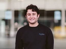 A person with curly hair smiling, wearing a black sweater with "Figma" written on it, standing indoors.