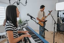 A young woman plays the keyboard and sings into a microphone while a man plays an electric guitar in a bright room.
