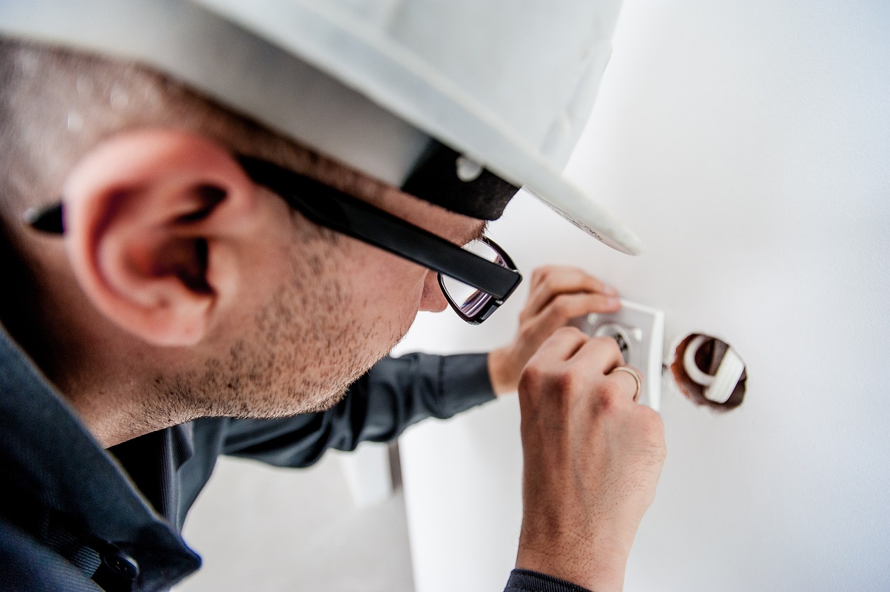 **Alt text:** A person wearing a hard hat and glasses is installing or repairing a power outlet on a wall.