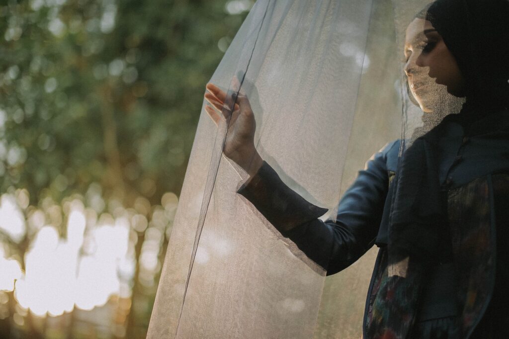 **Alt text:** A person wearing a hijab is softly touching a sheer curtain with sunlight filtering through the trees in the background.