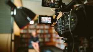 **Alt Text:** A video camera is set up in front of a person sitting in a blurred background, possibly a library, with a large light and shelves of books visible.